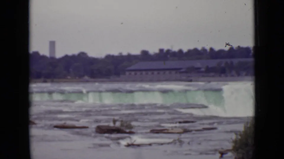 1940 Ontario home-movie frame showing water pouring over a broad edge, with scattered rocks, far shoreline buildings, and trees.