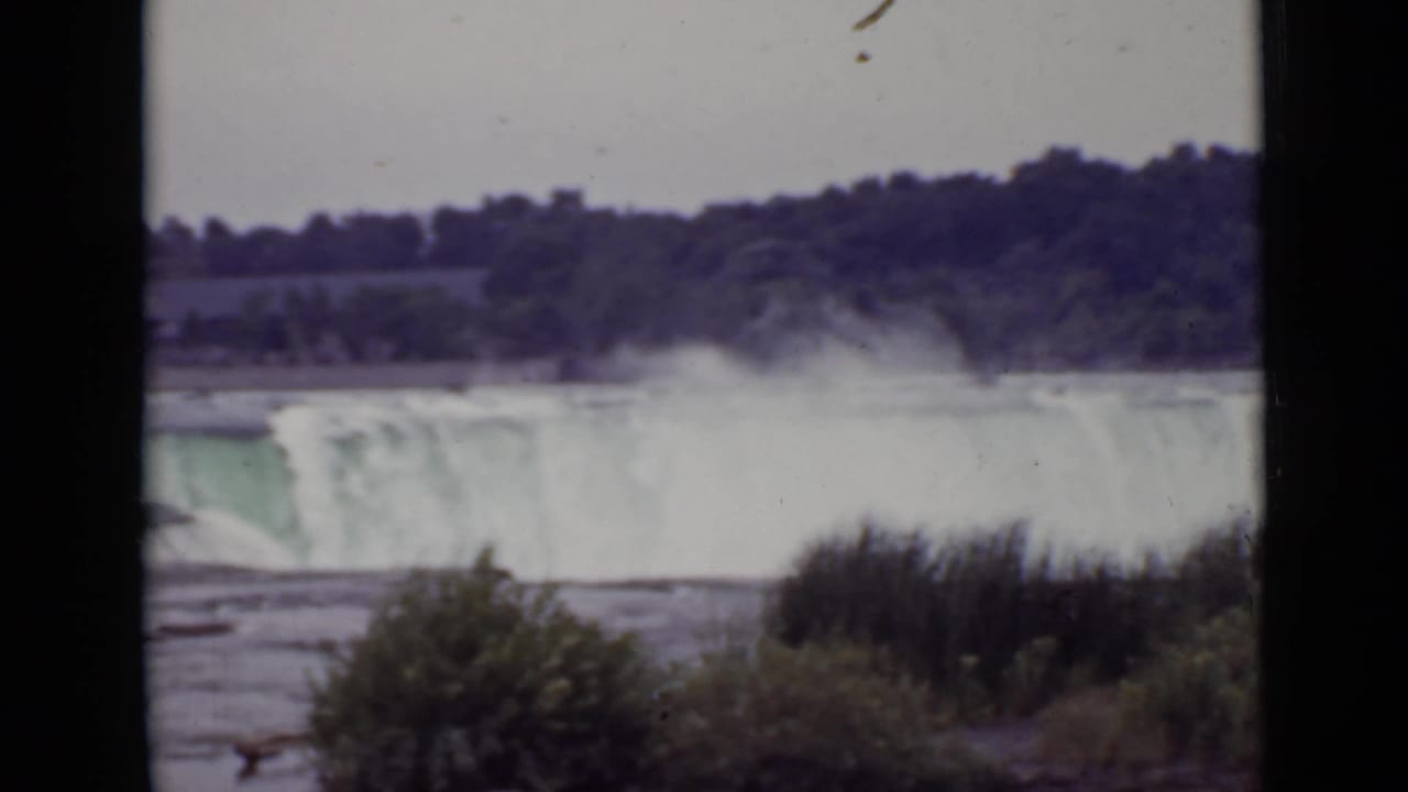 Wide view of Niagara Falls with mist, river, shrubs, and a tree-lined shore.