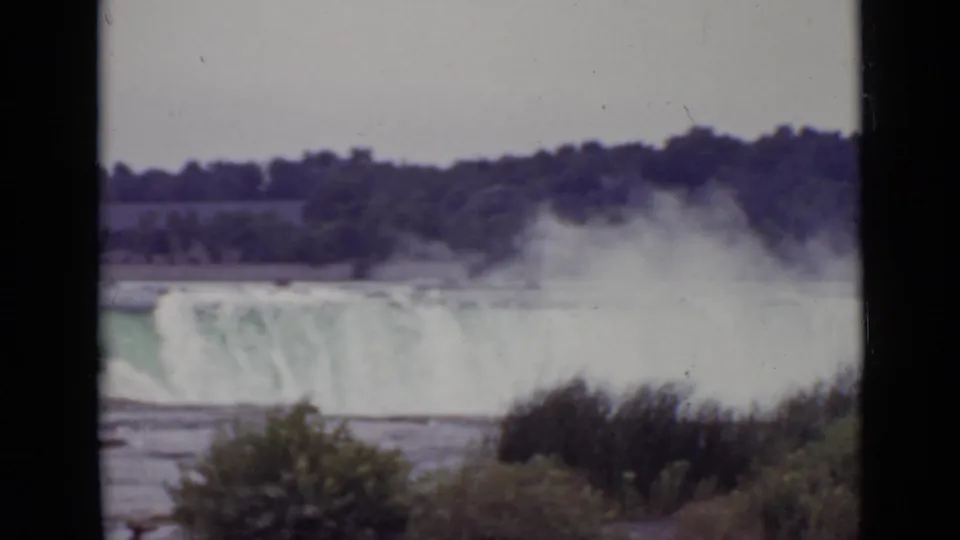 1940 Niagara Falls home-movie frame showing cascading water, heavy spray, foreground vegetation, and the wooded far shore.