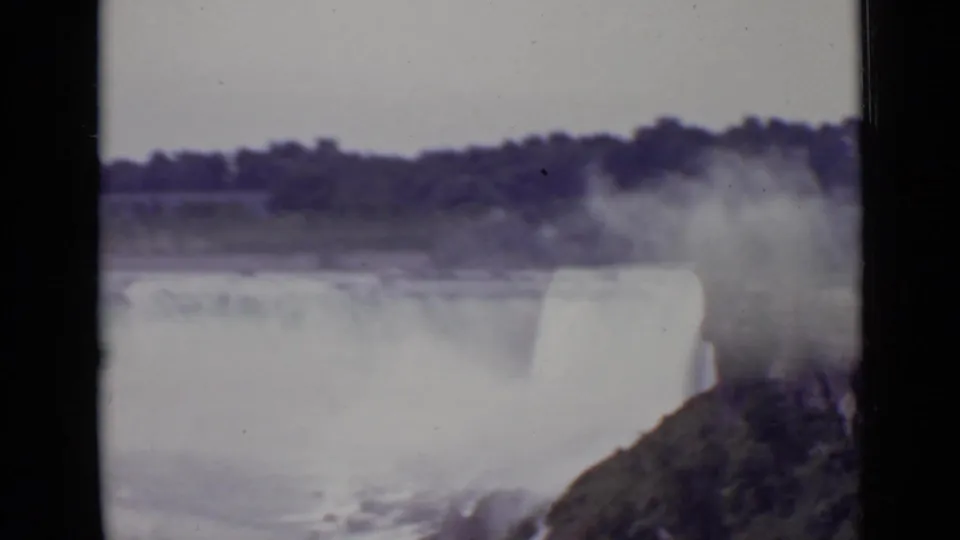 1940 8mm frame of Niagara Falls showing a closer waterfall drop, dense mist, turbulent water, and a vegetated rock edge.