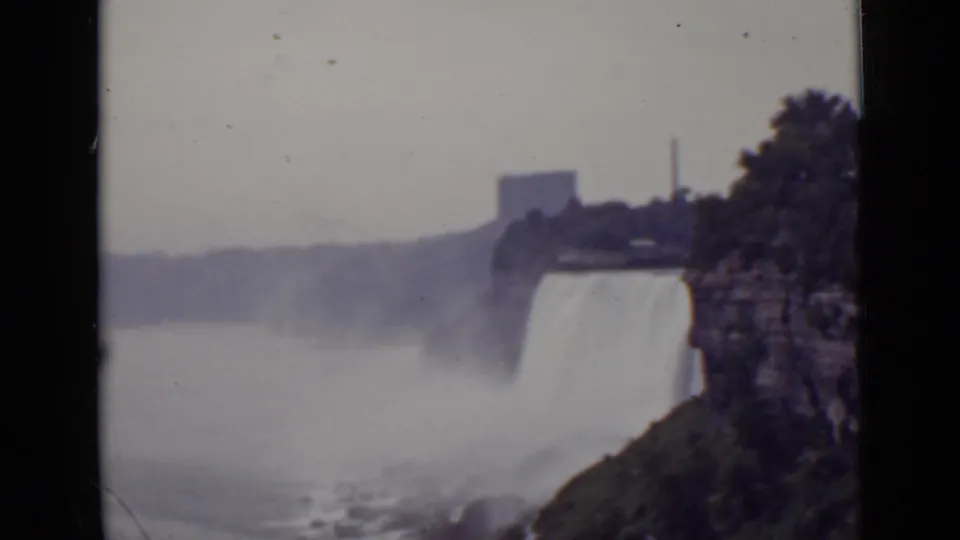 1940 Ontario view of Niagara Falls with a vertical drop, mist-filled gorge, rocky shoreline, trees, and distant buildings.
