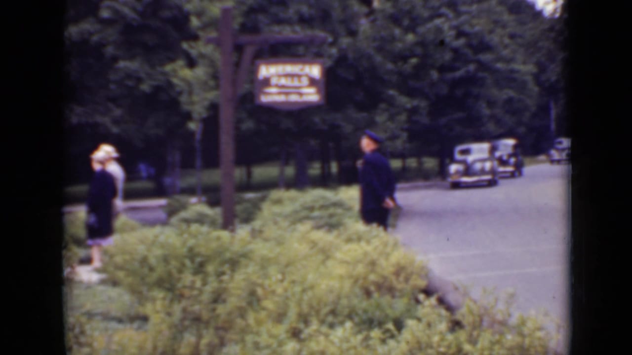 Traffic guard near an American Falls sign as pedestrians and cars appear along a tree-lined road.