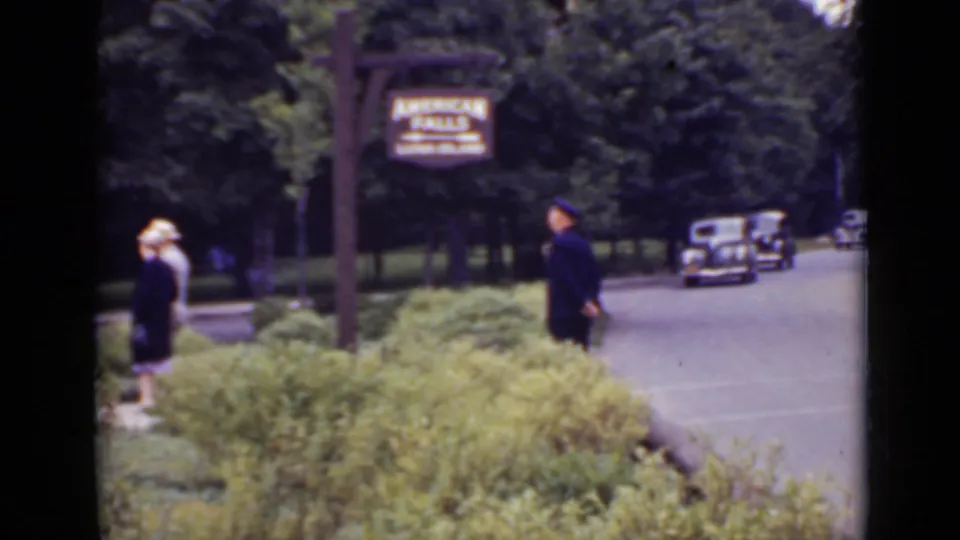 1940 Ontario home-movie frame showing a uniformed traffic guard near an American Falls sign with shrubs, trees, pedestrians, and period cars.