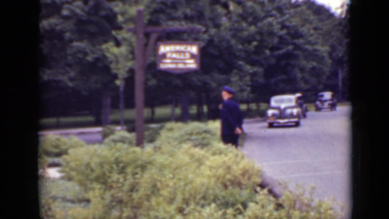 1940 Ontario frame with a uniformed guard beside the road under the American Falls sign while rounded automobiles approach through trees.
