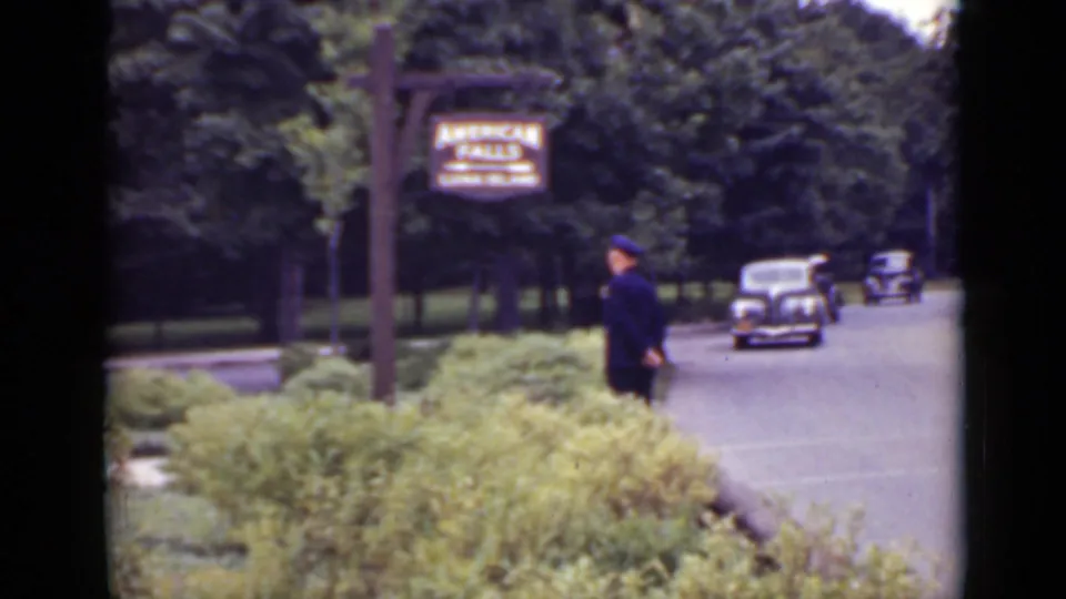 1940 Ontario frame with a uniformed guard beside the road under the American Falls sign while rounded automobiles approach through trees.