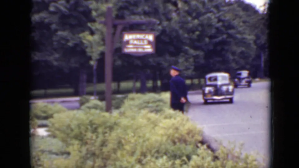 1940 Ontario tourist-road frame showing a dark-uniformed guard, American Falls sign, manicured shrubs, trees, and a passing black automobile.