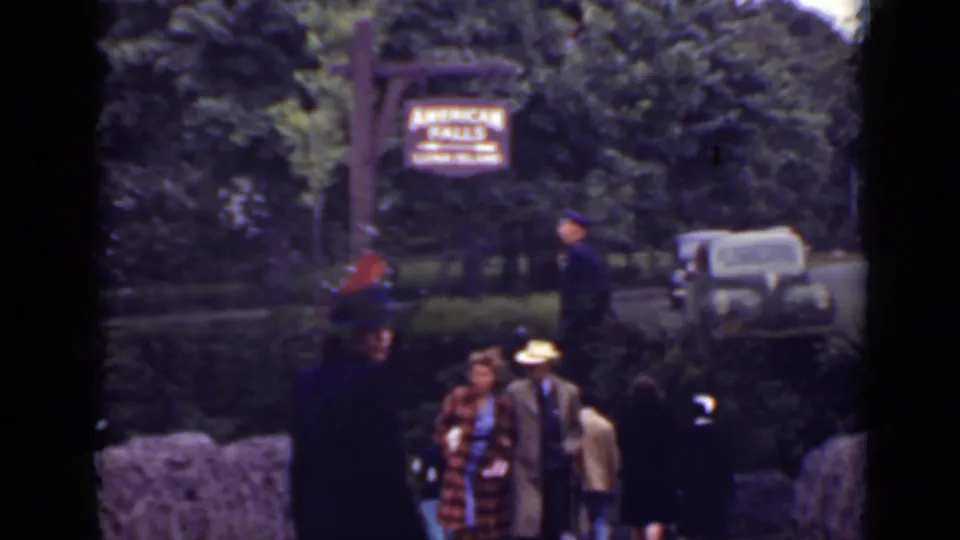 1940 Ontario home-movie frame with tourists in period hats, a stone wall, uniformed guard, car, trees, and the American Falls sign.