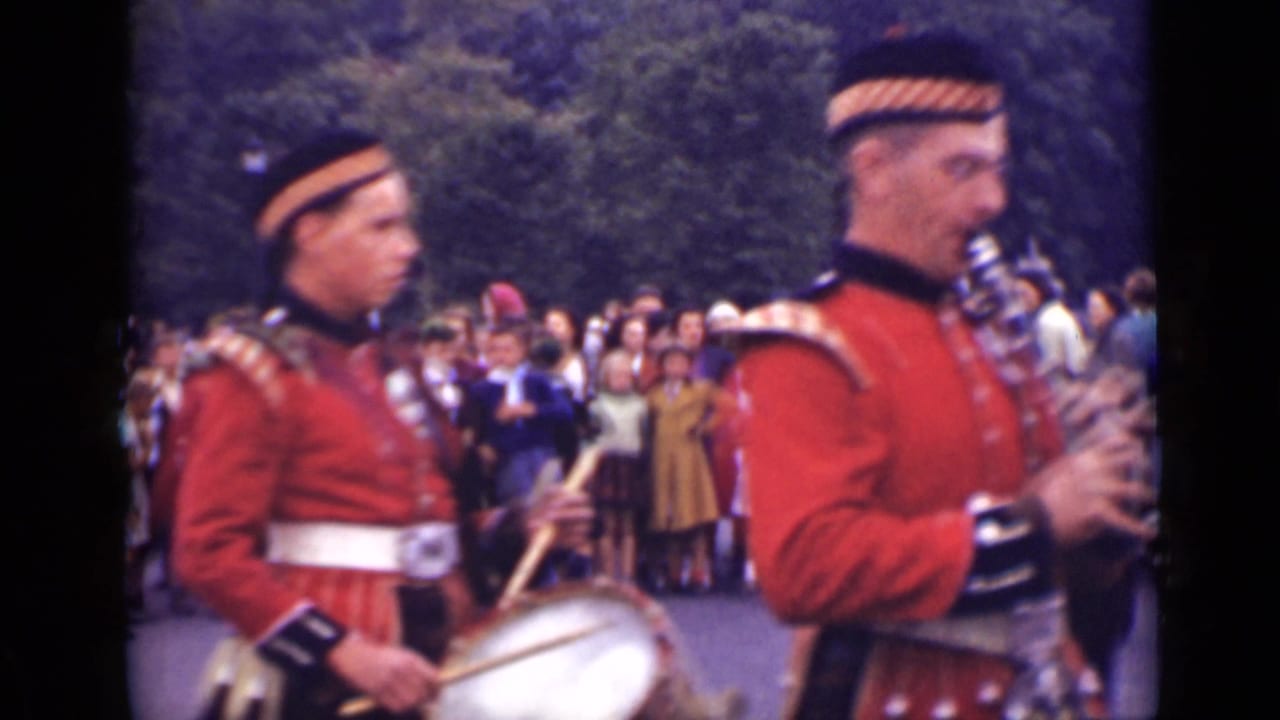Two uniformed band members in red jackets play drum and bagpipes before a crowd.