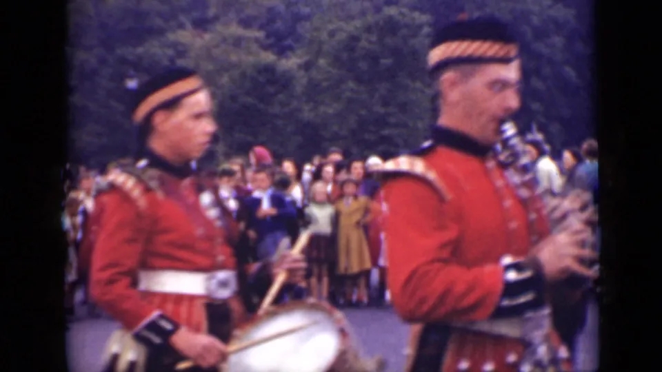 In 1940s Ontario home-movie footage, two Scottish-style band members in red tunics perform with drum and bagpipes as spectators watch.
