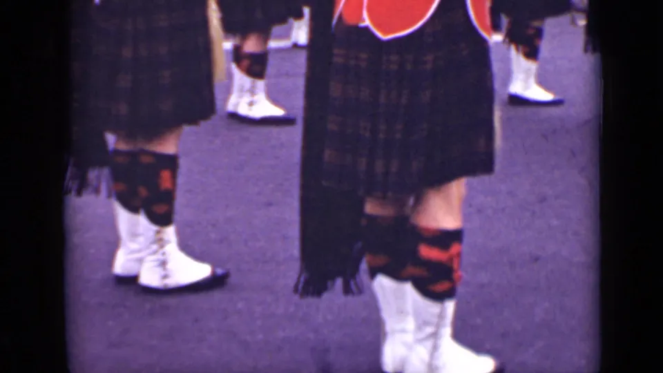 Close-up detail of Scottish-style parade dress, showing tartan kilts, patterned hose, and white gaiters on a street surface.