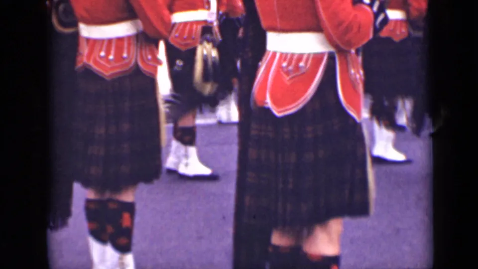 Kilted band members in red tunics and white belts stand in formation, with sporrans and tartan fabric visible from behind.