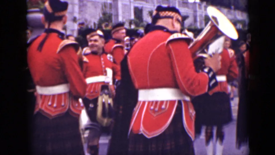 Scottish-style military band members in red jackets gather near spectators, one holding a large brass instrument during the Ontario parade scene.