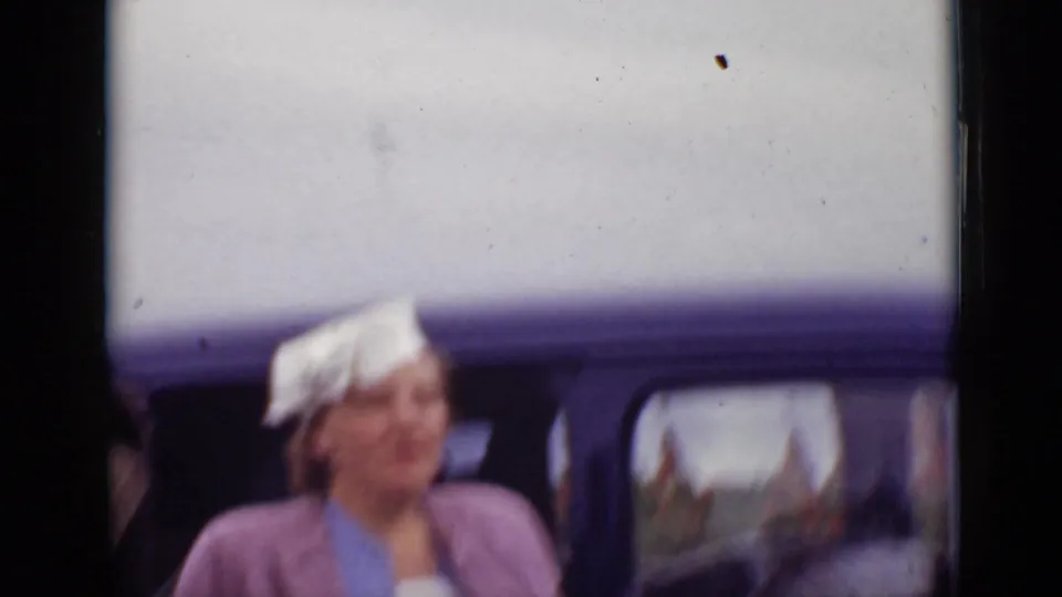 1940 Ontario frame with a woman in a white hat and pink outerwear beside a dark car in a crowded outdoor area.