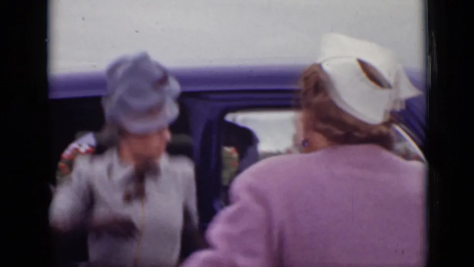 Two women in hats stand beside a dark car in 1940 Ontario, one wearing grey and the other pink outerwear.