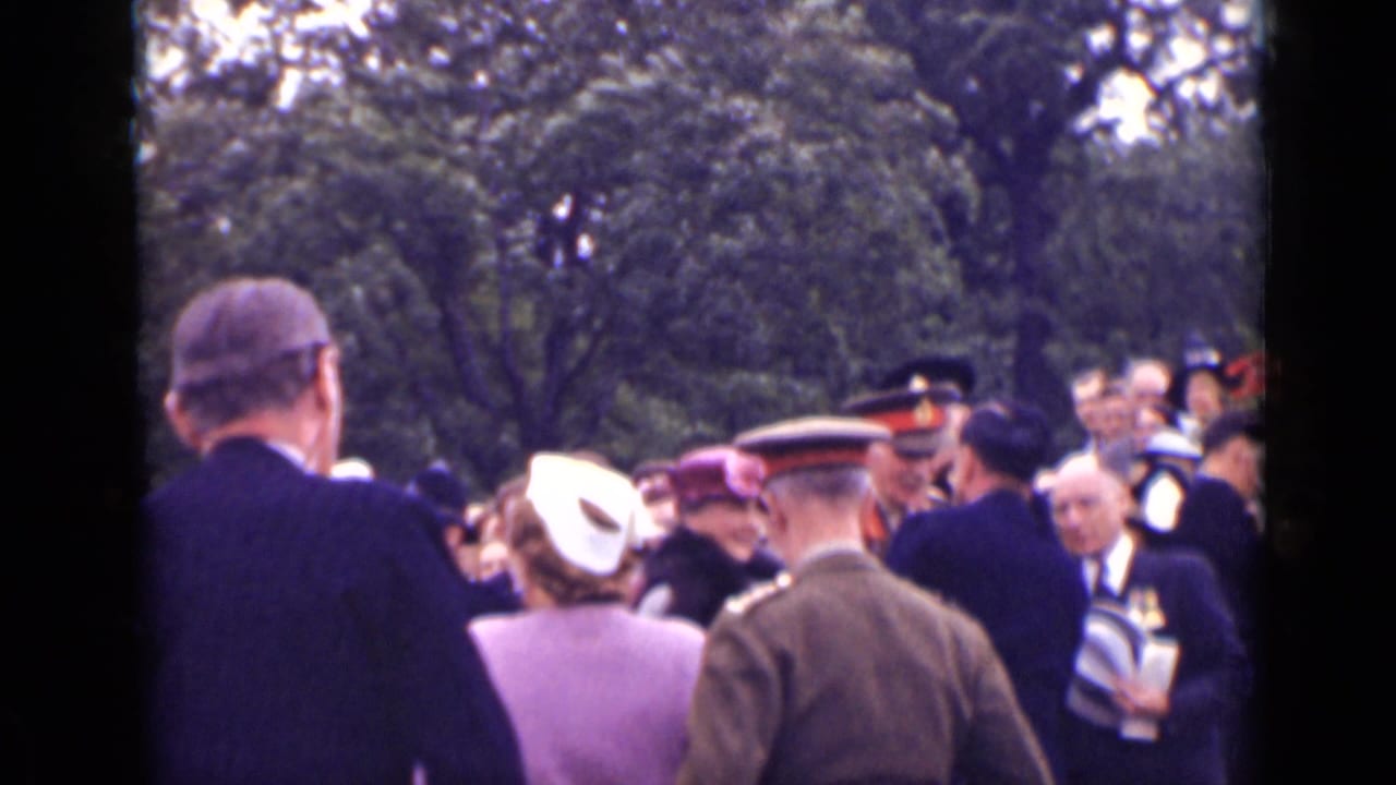 Outdoor crowd with civilians and uniformed men gathered under trees.