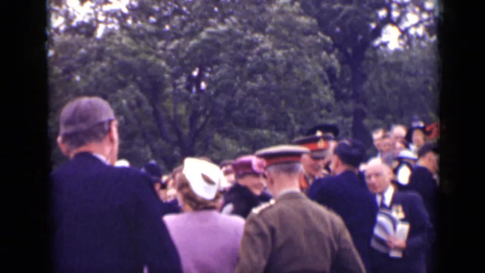 1940 Ontario home-movie frame showing a public outdoor crowd with hats, formal clothing, and several uniformed men under trees.