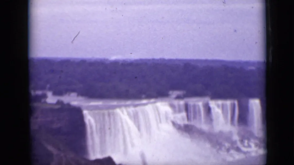 1940 Ontario 8mm frame showing Niagara Falls from a distant tourist viewpoint, with cascading water, mist, river, and far shoreline.