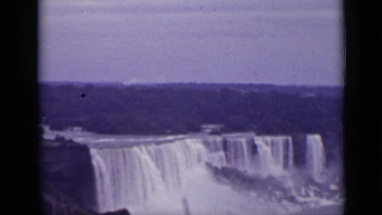 Wide view of Niagara Falls with mist, river water, and a tree-lined horizon.