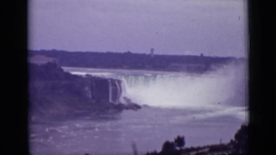 1940 Ontario frame showing Niagara Falls from a side angle, with a large mist cloud, river surface, distant shore, and foreground foliage.