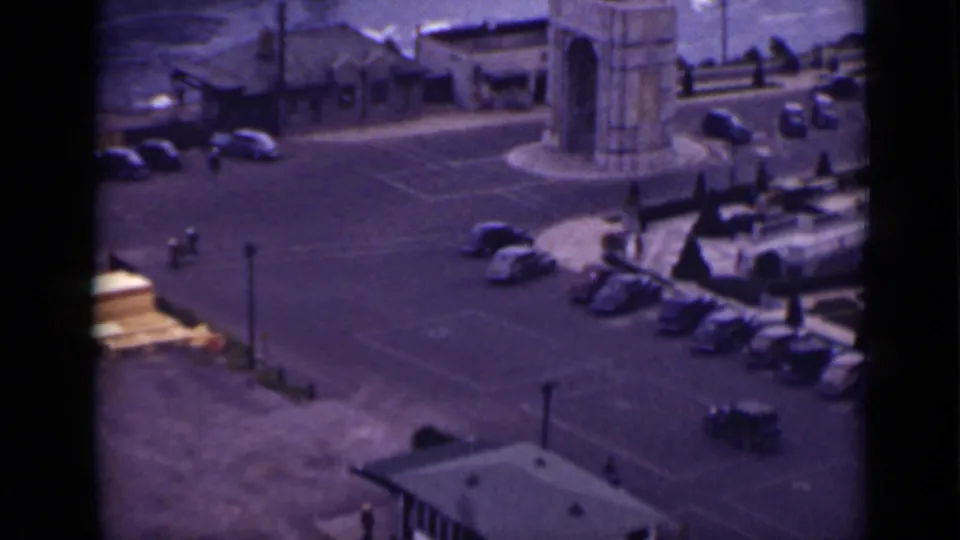 1940 Ontario Niagara Falls home-movie frame showing a parking lot, parked cars, a stone arch, railings, and water beyond.