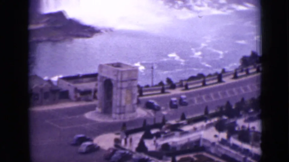 1940 Ontario Niagara Falls frame looking past the arch and parked cars toward turbulent water and heavy mist.