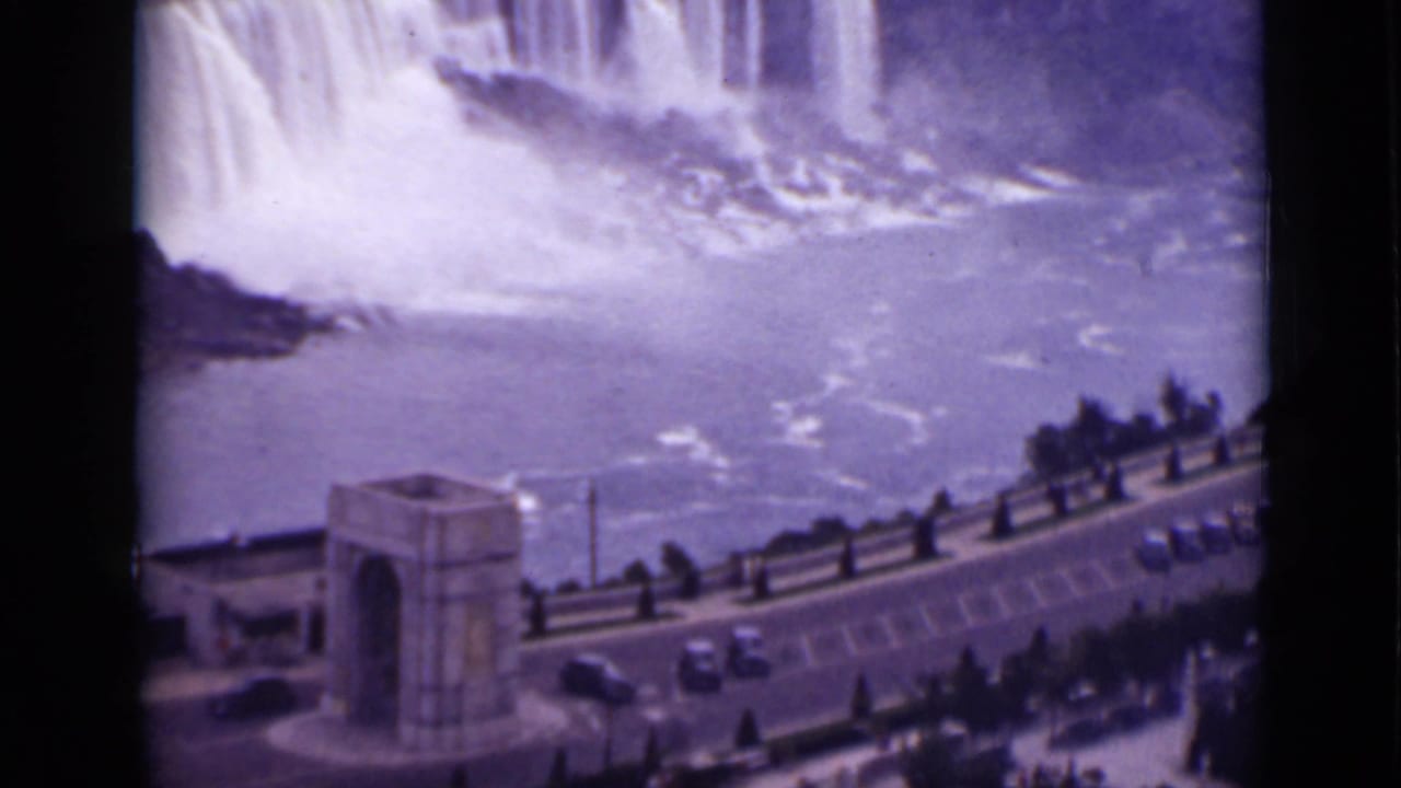Wide view of Niagara Falls with mist, river, overlook road, stone archway, and parked cars.