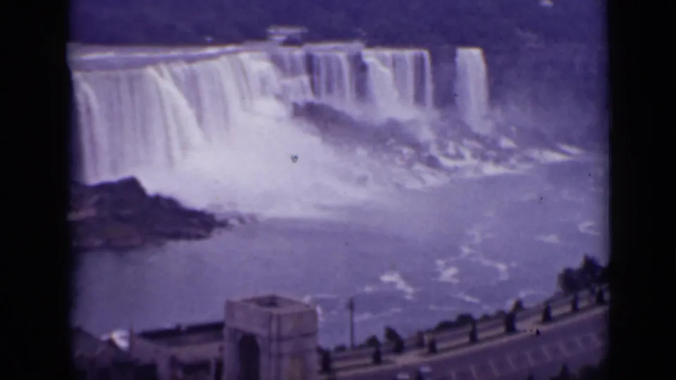 1940 Ontario travel frame of Niagara Falls, with cascading water, river mist, stone overlook structure, railing, and cars below.