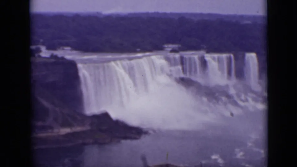 1940 Ontario 8mm frame showing Niagara Falls across the river with heavy spray, rocky foreground, and a dark treeline beyond.