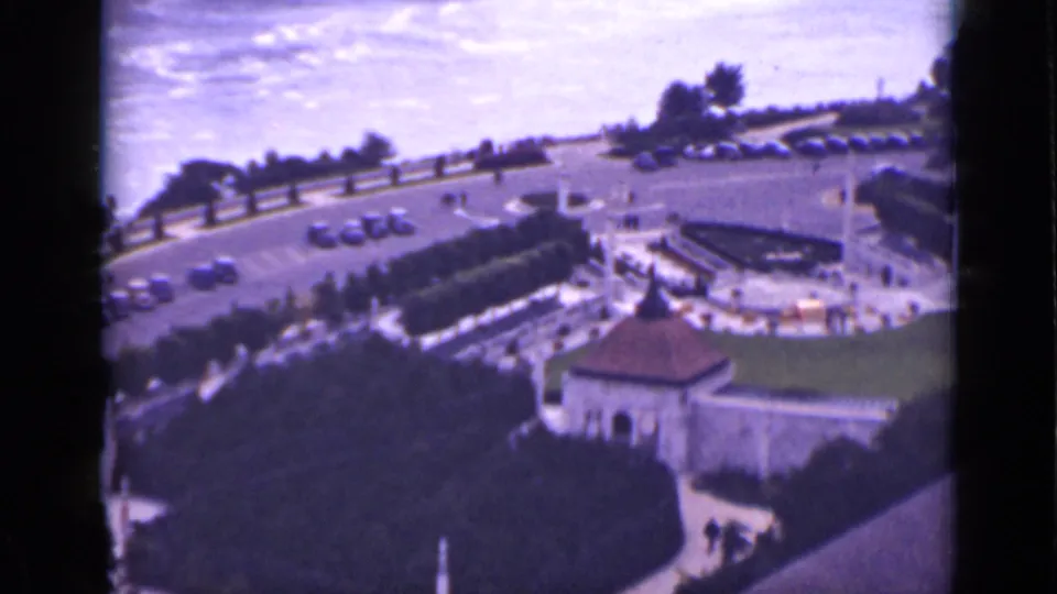 1940 8mm elevated view of the Niagara Falls visitor grounds in Ontario, with cars, pathways, trees, lawn, and distant water.