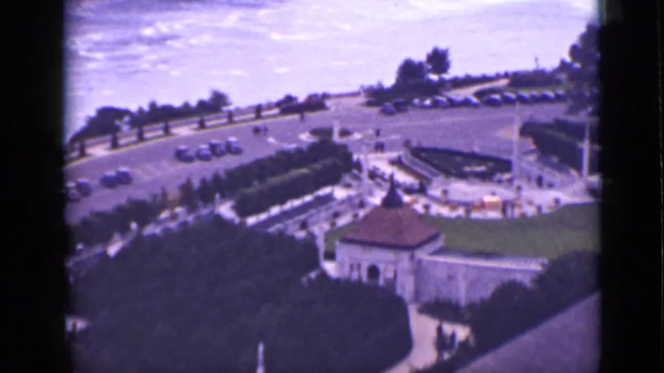 1940 8mm view over Niagara Falls visitor grounds in Ontario, showing parked cars, pedestrians, paths, trees, and small structures.