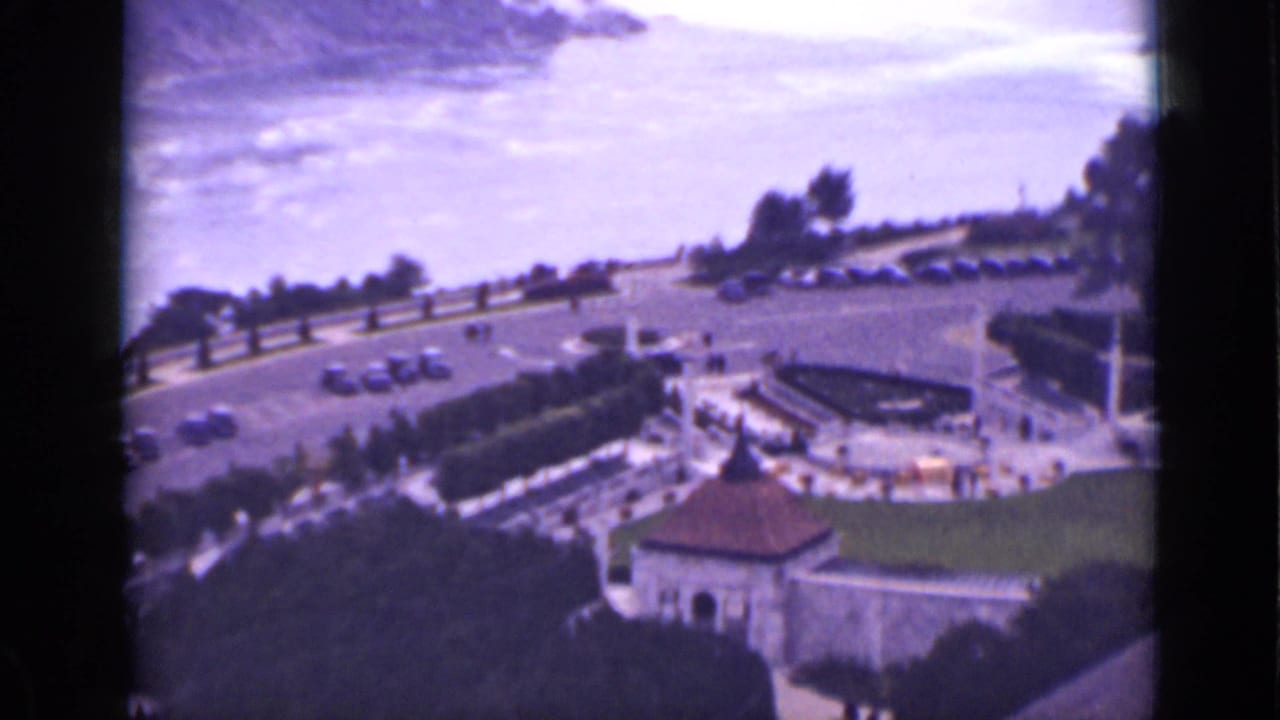 1940s Ontario home-movie view over Niagara Falls with a curved road, parked cars, landscaped grounds, and a small stone building.