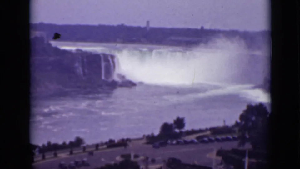 Faded 8mm view across Niagara Falls from Ontario, with mist, river water, a roadside edge, parked cars, and trees below.
