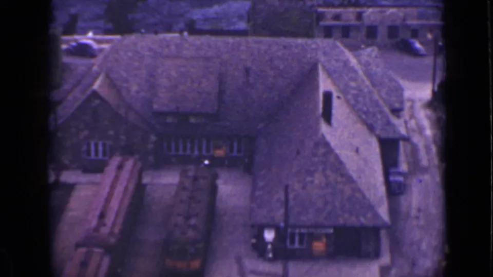 1940 Ontario home-movie frame with a distant elevated look over a steep-roofed building, vehicles, and adjacent ground.