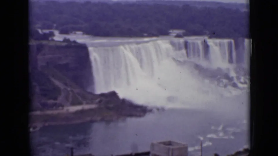 1940 Ontario home-movie frame showing Niagara Falls from a distant overlook, with mist, river water, and shoreline structures.