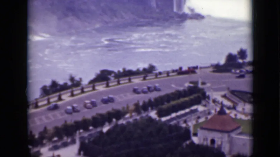 1940 Ontario 8mm frame showing the road below Niagara Falls, with cars, trees, a small roofline, water, and waterfall spray in the distance.