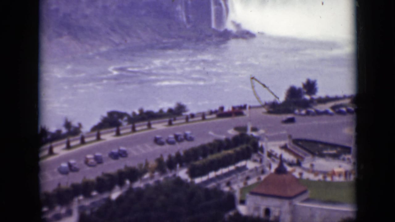 Elevated view of Niagara Falls with a curved road, cars, hedges, and overlook buildings below.