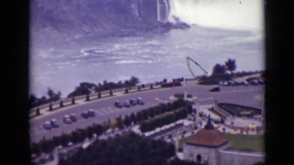 1940 Ontario home-movie frame showing Niagara Falls above a curved tourist road with cars, hedges, and small overlook structures.