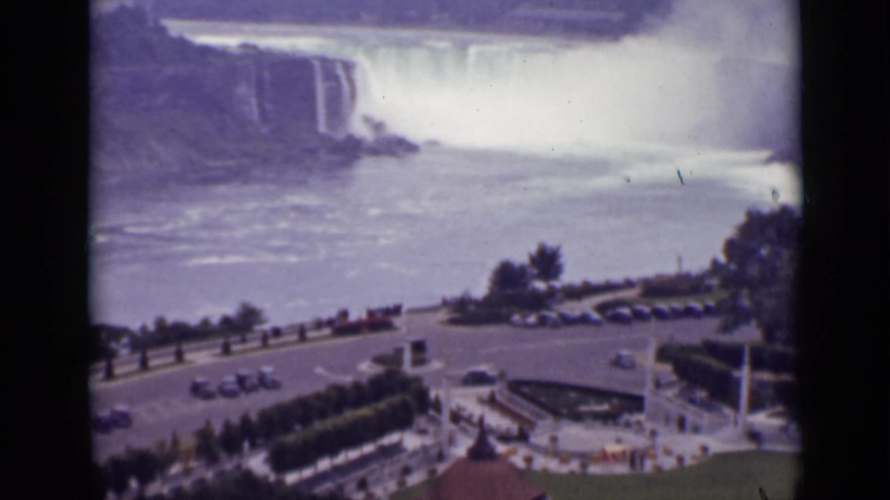 Niagara Falls and mist above a roadside overlook with several cars and landscaped paths.