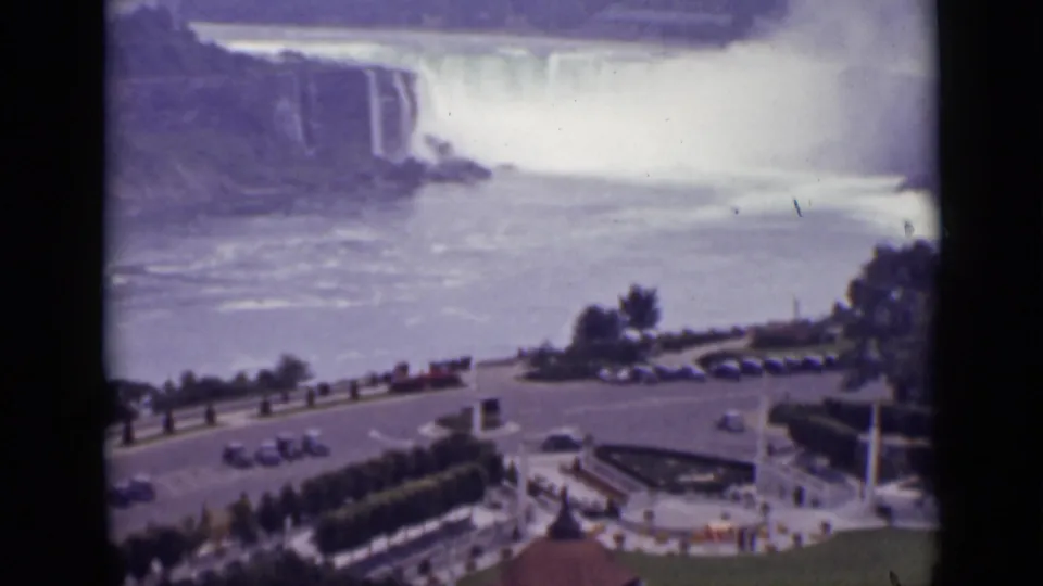Elevated 1940 Ontario view of Niagara Falls with mist, a roadside overlook, parked cars, and manicured landscaping below.