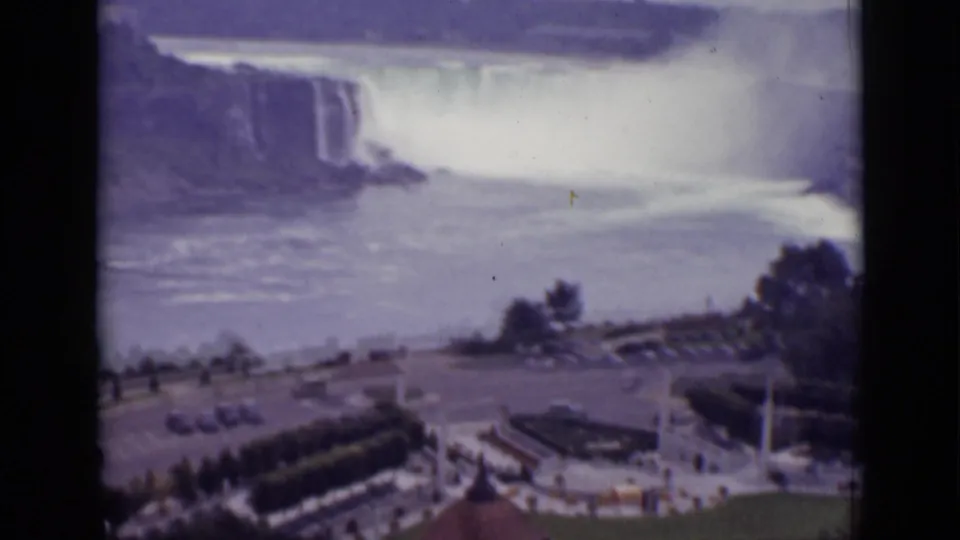 Soft 8mm frame of Niagara Falls in Ontario, with road traffic, trees, railings, and visitor-area structures in the foreground.