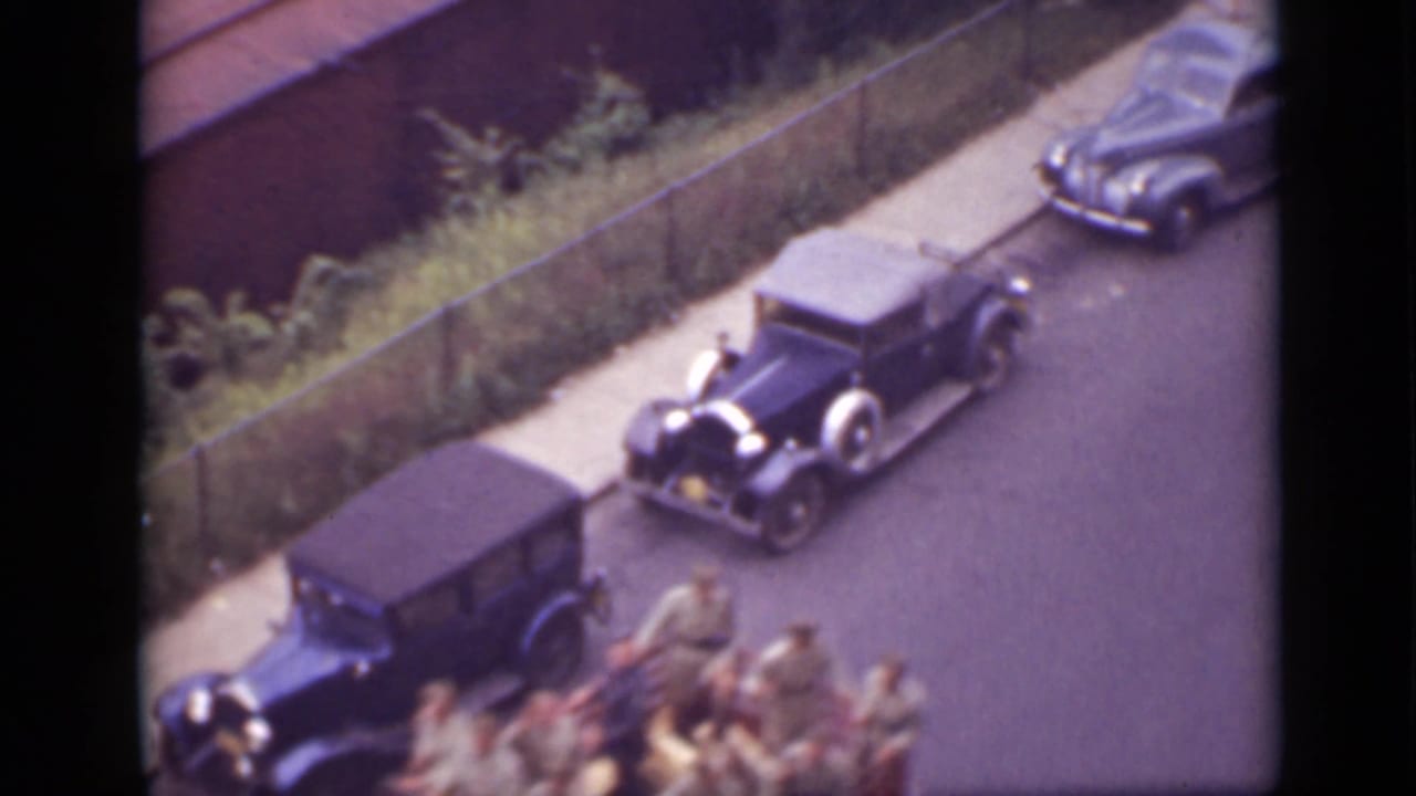 High-angle view of vintage cars parked along a street with people in a vehicle at the lower edge.