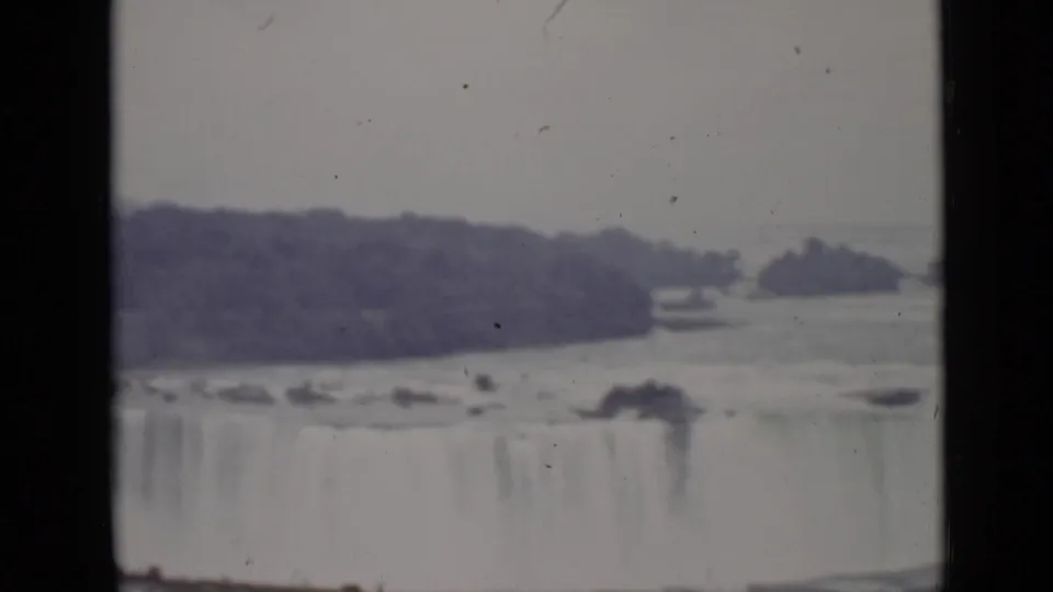 Distant 1940 Ontario view of Niagara Falls, showing the broad cascade, river surface, mist, and dark wooded landforms.