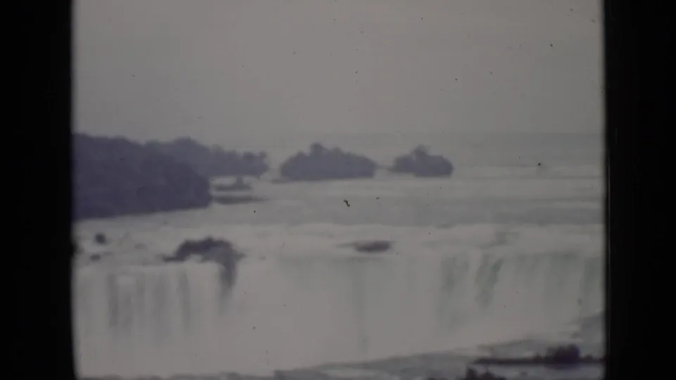 1940 home-movie frame showing Horseshoe Falls from Ontario, with pale water spilling over the brink and wooded islands across the river.