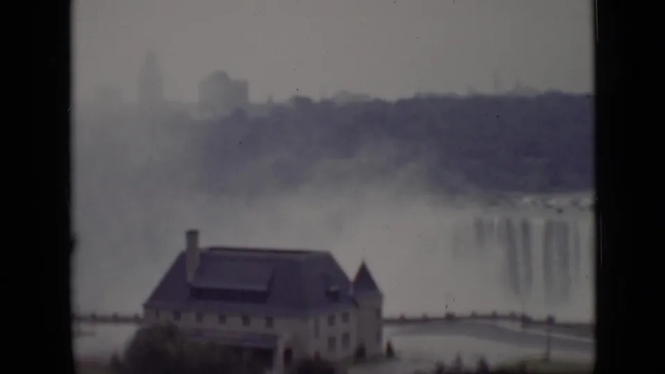 1940 Ontario 8mm frame with a large riverside building in the foreground and mist rising from Niagara Falls behind it.