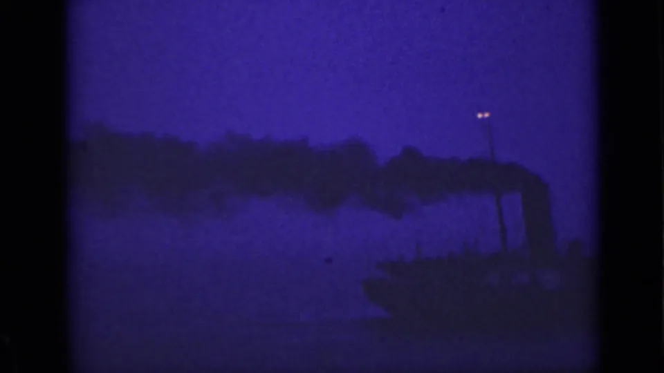 1940 Ontario home-movie frame with a dark smokestack and smoke plume silhouetted against low-light water and sky.