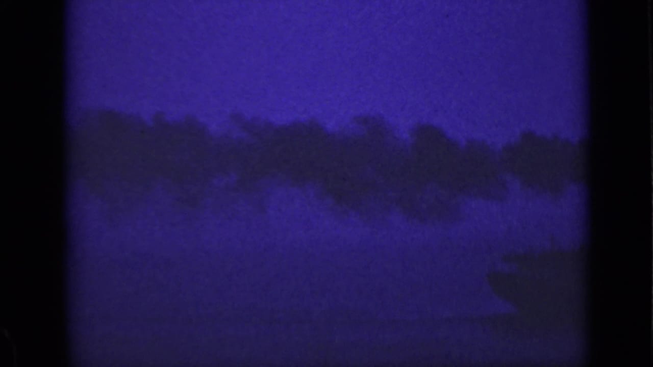 Broad smoke plume stretches across a dark blue night scene over water.