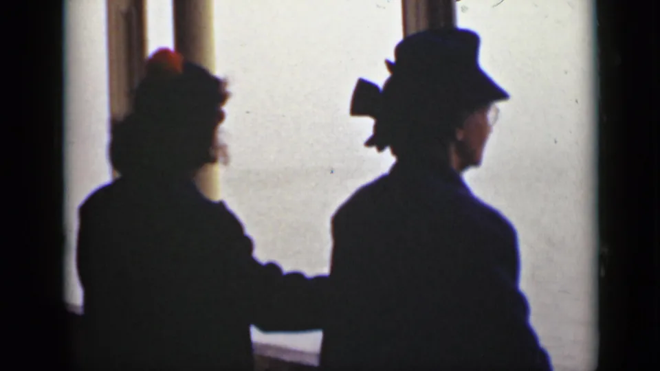 Two women in period coats and hats stand in silhouette by a large bright window in 1940 Ontario home-movie footage.