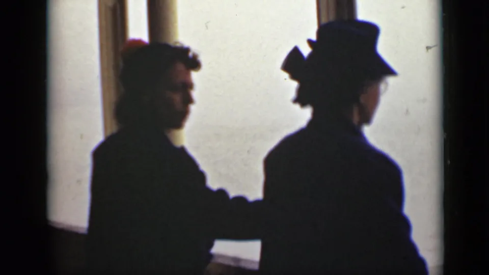 In backlit 8mm footage, two women in hats stand by a window, with one figure partly turned toward the other.