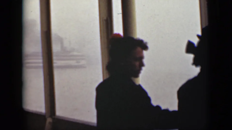 The women remain near the window as a faint ship or shoreline shape appears across the bright water outside.