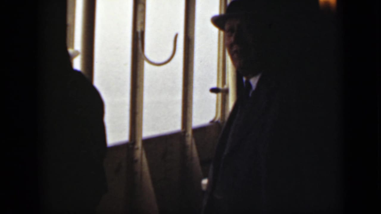 Man in a dark fedora standing beside bright vessel windows.